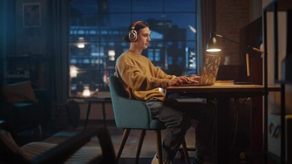 Wall Mural - Young Handsome Man Working from Home on Laptop Computer in Stylish Loft Apartment in the Evening. Creative Person Checking Social Media, Browsing Internet. Urban City View from Big Window.