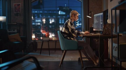 Fototapete - Young Woman Writing Code on Desktop Computer in Stylish Loft Apartment in the Evening. Creative Female Wearing Cozy Clothes, Working from Home on Software Development. Urban City View from Big Window.
