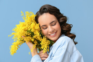 Beautiful young woman with bouquet of mimosa flowers on blue background