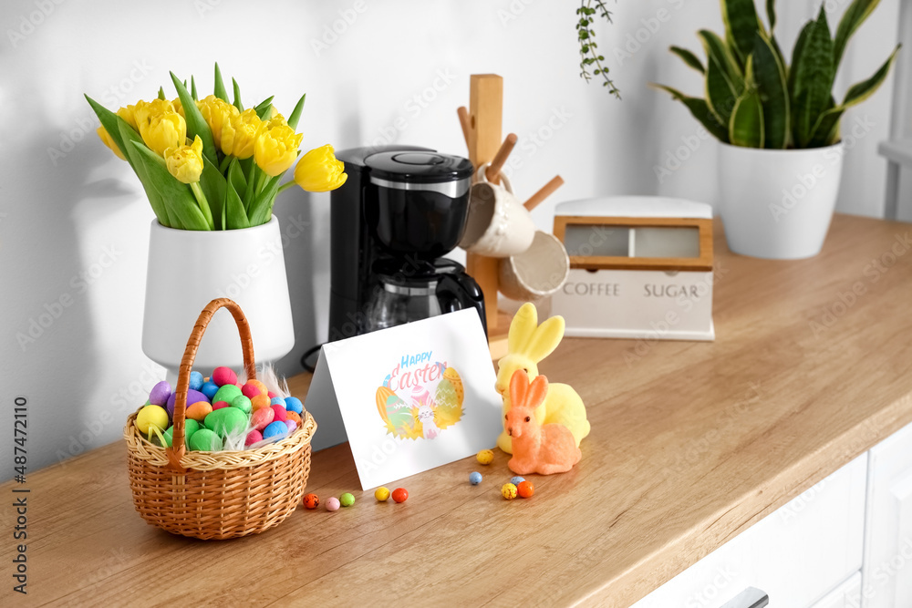 Basket with Easter eggs, rabbits, greeting card and tulips on counter near light wall