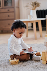 Wall Mural - Concentrated curly black boy sitting on carpet in living room and watching cartoons on tablet