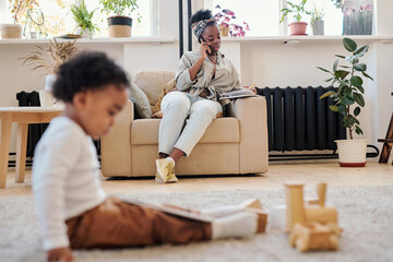 Wall Mural - Busy young African-American mother in headscarf sitting in armchair and talking by phone while working during maternity leave