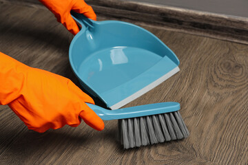 Woman in gloves sweeping wooden floor with plastic whisk broom and dustpan indoors, closeup