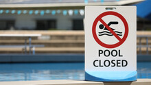 A Bright White Sign At A Local Outdoor Pool Facility Saying Pool Is Closed. No Entry To Water. Clear Clean Blue Water At Public Baths And No Swimming Symbol.