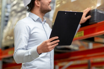 Wall Mural - logistic business, shipment and people concept - close up of businessman in helmet with clipboard checking goods at warehouse