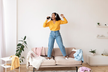 Wall Mural - Cool black woman in headphones using smartphone as mic, standing on couch, singing songs, checking mobile music app