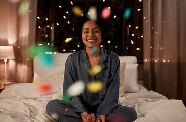 Sticker - holidays, christmas and people concept - happy smiling woman in pajamas sitting in bed over colorful confetti at night at home