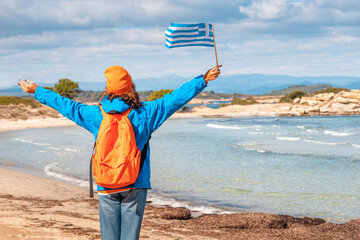 Wall Mural - A happy woman wearing jacket and hat with a Greek flag on the beach of the resort. Off and low season vacation and tourism concept