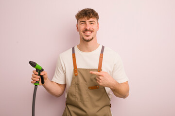 Wall Mural - young cool man smiling cheerfully, feeling happy and pointing to the side. gardener and hose concept