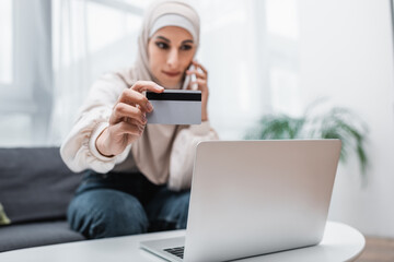 Wall Mural - selective focus of credit card in hand of blurred arabian woman calling on smartphone near laptop.