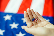 Close Up Of Young Woman Holding United State Of America Pin In Her Hand. American Flag On The Background. USA Patriotism Concept.