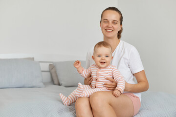 Wall Mural - Portrait of positive delighted young adult mother wearing white casual t shirt sitting on bed in bedroom with toddler baby, female holding her kid and looking at camera with optimistic emotions.