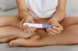 Closeup portrait of female hand holding positive pregnancy test while sitting on bed with crossed legs. Motherhood, pregnancy, birth control.