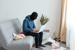 Portrait of man wearing blue hoodie and black robber mask, breaking flat, sitting on sofa and opening laptop, stealing modern technique, finding important files on notebook.