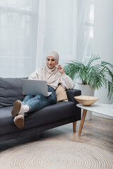 Wall Mural - cheerful arabian woman in hijab eating popcorn and watching film on laptop on couch.