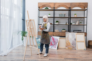 Wall Mural - full length view of muslim woman drawing on easel near painted pictures in home studio.