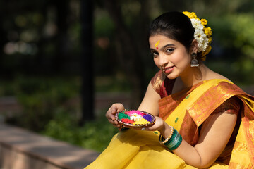 Wall Mural - Beautiful and young girl or woman in yellow saree seating in garden with a decorated plate of color or colour or gulal or abeer or Holi powder to celebrate holi festival of colors