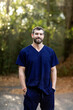 A doctor with dark hair and a beard in navy blue scrubs standing outside in a natural green environment