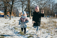 Two Sisters Have Fun And Actively Spend Time In The Park In Winter, They Joyfully Run Together In The Snow.