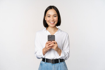 Wall Mural - Stylish modern asian girl using mobile phone application, chatting on cellphone and smiling, standing in white blouse against studio background