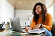 © Prostock-studio - Black woman sitting at desk, using computer writing in notebook
