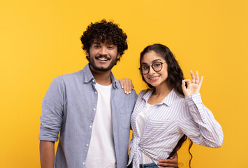 Wall Mural - Positive indian spouses embracing and lady showing okay gesture over yellow studio background