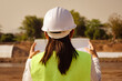 © Pornpimon - Asian female engineer wearing a helmet and safety vest works and looks at the blueprints for construction plan and design details of the dam.