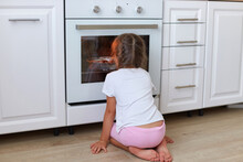 Back View Of Little Girl Sitting Near Oven In Kitchen And Looking Inside Through Glass, Curious Impatient Female Child Waiting For Cookies Baking In Stove, Enjoying Homemade Pastry.