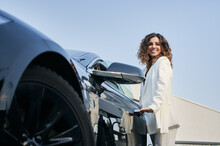 Bottom View Of Happy Smiling Business Girl With Curly Hair Standing Outside Car And Easily Opening Door Of Her Auto. Beautiful Woman Holding Handle On Car Door On The Background Of Blue Sky.