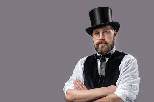 portrait of confident bearded magician wearing eyeglasses, bow tie and top hat posing in studio with