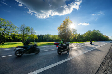 three motion blurred motorcycles riding on the asphalt road along a spring meadow in the woods at su