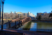 A Rust Colored Iron Bridge Over A Green Lake In The Park With Buildings In The City Skyline Surrounded By Bare Winter Trees And Lush Green Trees And Plants With Blue Sky In Downtown Atlanta Georgia