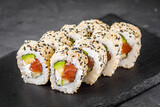 Fototapeta Kuchnia - appetizing california sushi roll with cheese avocado cucumber and salmon in sesame seeds on a black stone plate