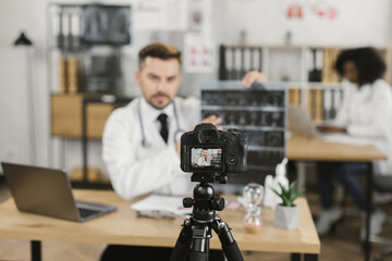 Competent male physician in uniform recording video blog while examining x ray scan at hospital room. Focus on digital camera fixed on tripod.