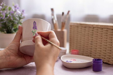 Female hands paints with a brush a sprig of a lavender flower, on a white ceramic pot