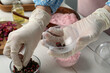 Woman in gloves filling bath bomb mold with flower buds at white table, closeup