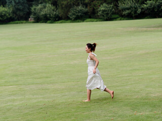 Wall Mural - Portrait of young Asian woman running on grass field with barefoot in forest park, beautiful Chinese girl in white dress enjoy her carefree time in sunny summer day, full length shot.