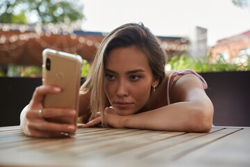 young asian pretty woman with long hair lays her head on her arms and table, holding phone in hand. beautiful pensive melancholic lady laying at cafe, using smartphone. lifestyle portrait