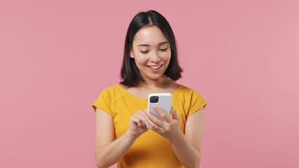 Wall Mural - Dreamful minded smart young woman of Asian ethnicity 20s wears yellow t-shirt hold using mobile cell phone swipe typing browsing chatting isolated on plain pastel light pink background studio portrait