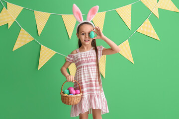 Little girl with bunny ears and basket closing eye with Easter egg on green background