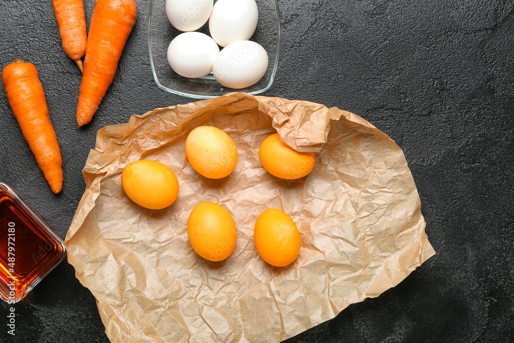 Parchment with Easter eggs, carrots and bowl with homemade natural dye on dark background