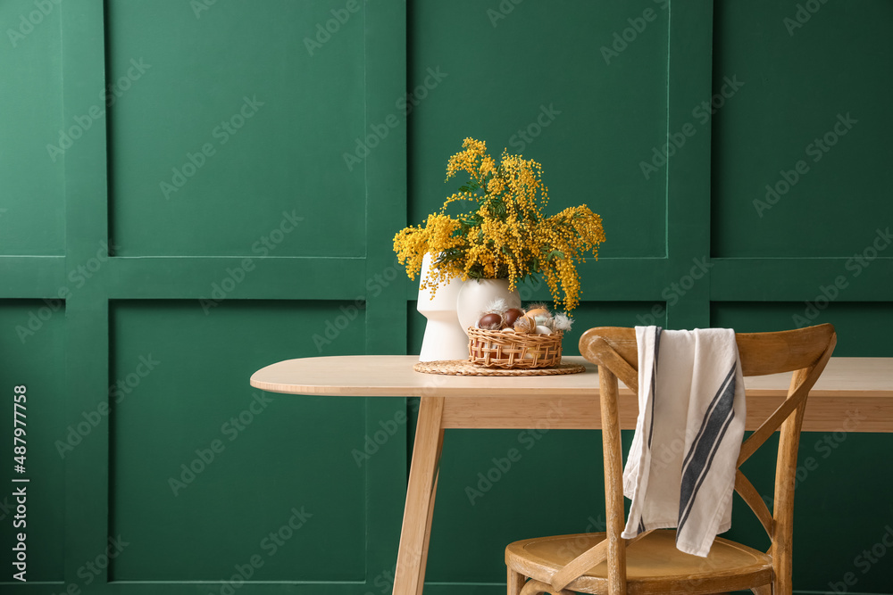 Vase with mimosa flowers, basket and Easter eggs on dining table near green wall
