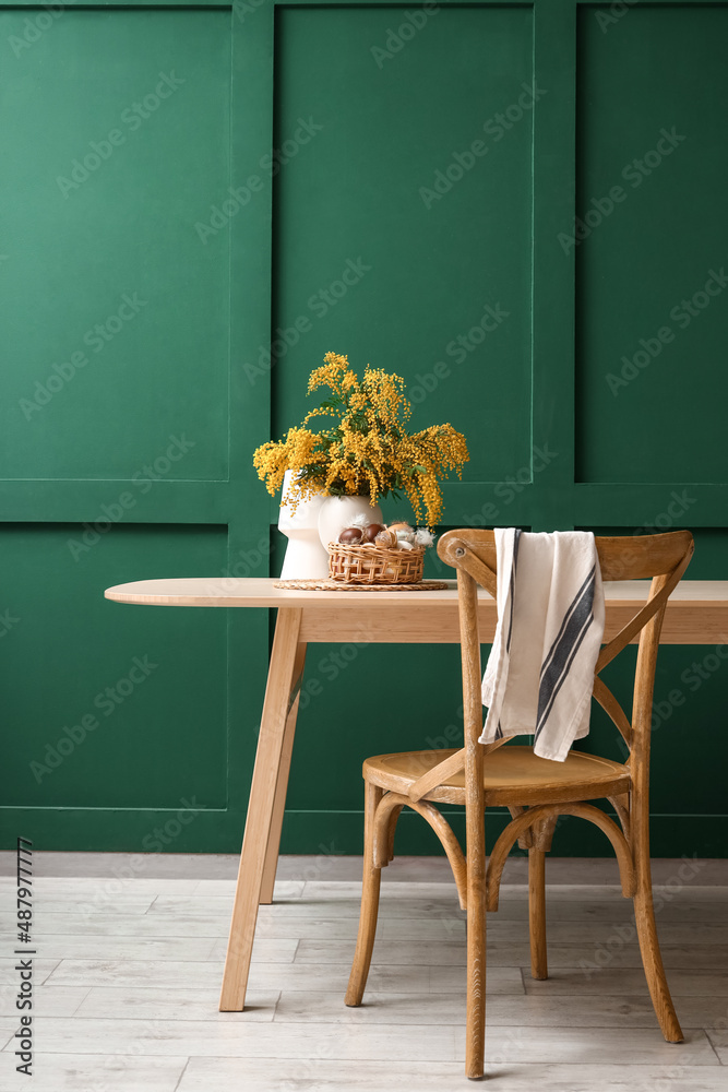Vase with mimosa flowers, basket and Easter eggs on dining table near green wall