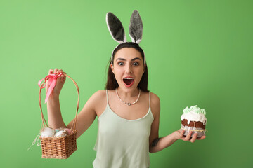 Surprised young woman with tasty Easter cake and eggs in basket on green background