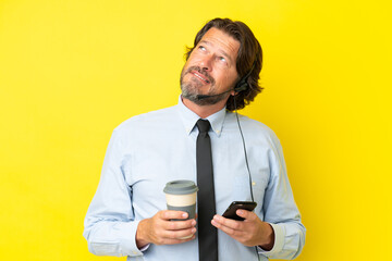 Telemarketer dutch man working with a headset isolated on yellow background holding coffee to take away and a mobile while thinking something