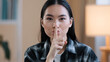 © Yuliia - Portrait asian ethnic korean woman point with finger near mouth Chinese Japanese girl makes hush quiet gesture showing secret sign asks silent confidential secrecy information looking at camera indoor