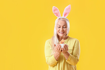 Wall Mural - Mature woman with bunny ears and toy rabbit on yellow background. Easter celebration