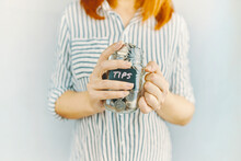 Glass Bottle With Silver Coins In Women's Hands. Sticker With Inscription On Money Jar. Redhead Girl N White And Black Striped Shirt Holds Her Tips Pot.