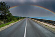 A rainbow with a thundercloud on the horizon of a highway. Rainbow in the storm. Asphalt pavement. Road markings on asphalt. Cloud horizon. Coastal road. Go on vacation. Summer season.