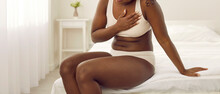 African American Woman Suffering From Heart Disease. Young Black Lady In White Underwear Suddenly Feels Pangs Of Pain, Sits Down On The Edge Of Her Bed And Touches Her Chest. Cardiac Pain Concept